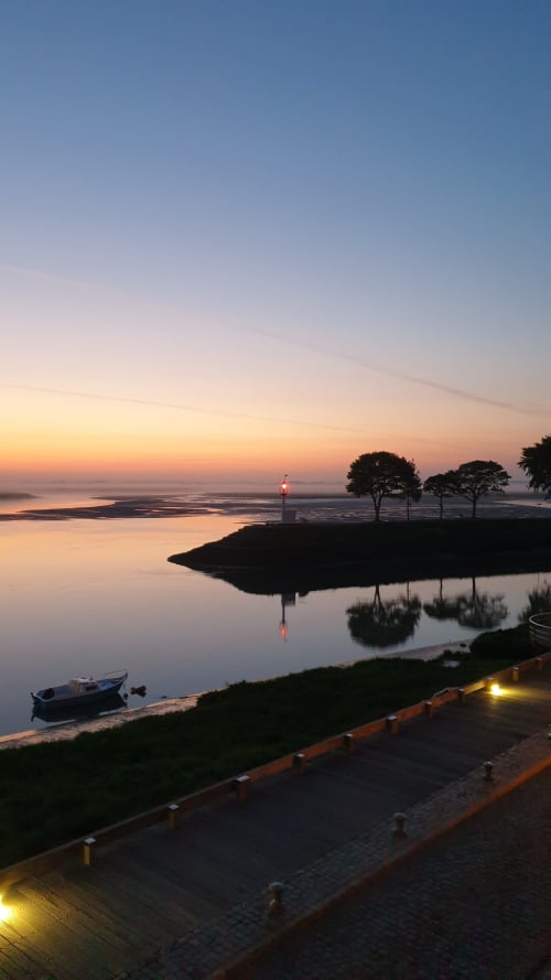 Vue du coucher de soleil sur le port de Saint Valery sur Somme avec phare, bateaux et arbres reflétés dans une eau calme