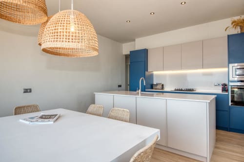 Modern kitchen with white cabinetry, blue accents, woven pendant light, dining table, and island with sink in Palafrugell