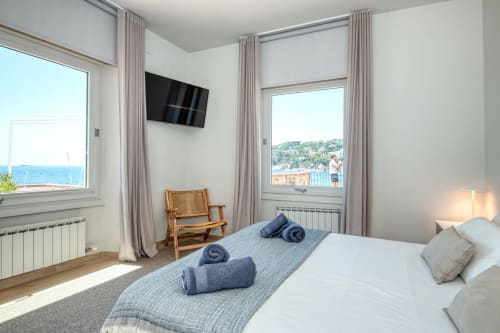 Modern bedroom with sea view at Casa dins el Mar, Palafrugell. Bed with blue towels, TV, wooden chair, windows overlooking