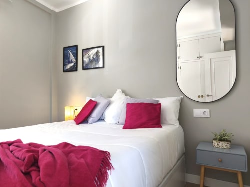 Dormitorio moderno en apartamento Holibai, Baiona con cama blanca, almohadas rojas, espejo redondo y obra de arte costera