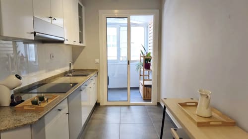 Cocina tipo galera moderna en apartamento de Baiona con armarios blancos, encimeras de granito y puertas de cristal que se abren a balcón con vistas a la ciudad