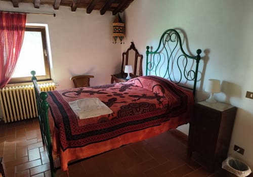 Camera rustica ad Agri-Bio Santa Palmira con letto in ferro battuto ornato, biancheria rossa con motivi e travi in legno a vista a Pomarance