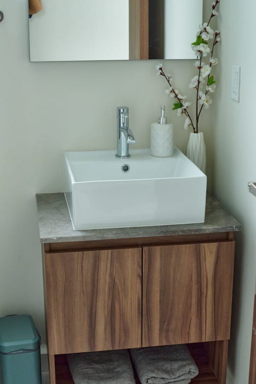 Moderno tocador de baño en apartamento Pares Homes CDMX con lavabo blanco, gabinete de madera y flores decorativas.
