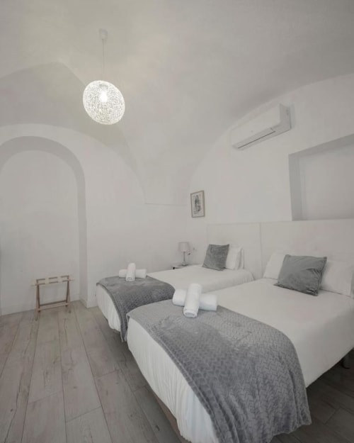 Dormitorio minimalista con dos camas en apartamento de Mérida, paredes blancas, ropa de cama gris, lámpara colgante y aire acondicionado