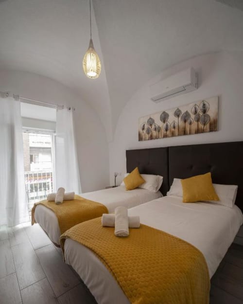 Dormitorio luminoso en Mérida con dos camas, detalles mostaza, lámpara colgante y balcón con vista a la ciudad