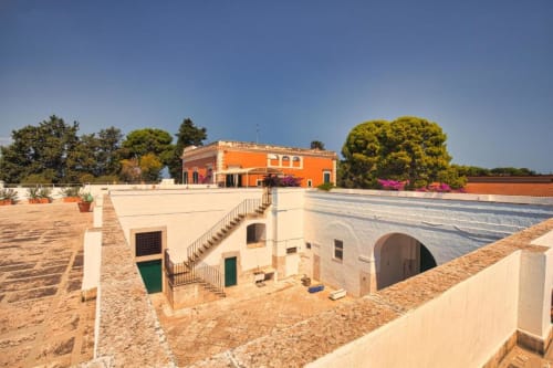 Corte della Villa Ceci ad Andria con edifici color crema, villa arancione, ingresso ad arco e alberi verdi lussureggianti sotto cielo azzurro