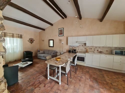 Cucina a pianta aperta e zona living con soffitto a travi, piastrelle in terracotta, mobili bianchi e tavolo da pranzo al Gîte Le