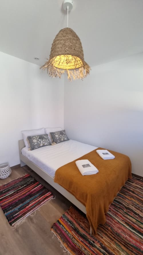 Quarto aconchegante na Casa da Oliveirinha em Leiria com luminária pendente em corda natural, cama branca e tapete listrado colorido.