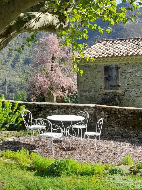 Patio du L'Amiradou BnB à Félines-sur-Rimandoule avec table et chaises en fer forgé blanc entourées de verdure.