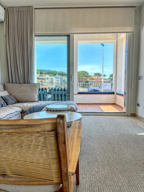 Bright hotel room with coastal views, bed, wooden chair, and terrace overlooking Palafrugell harbor, Spain