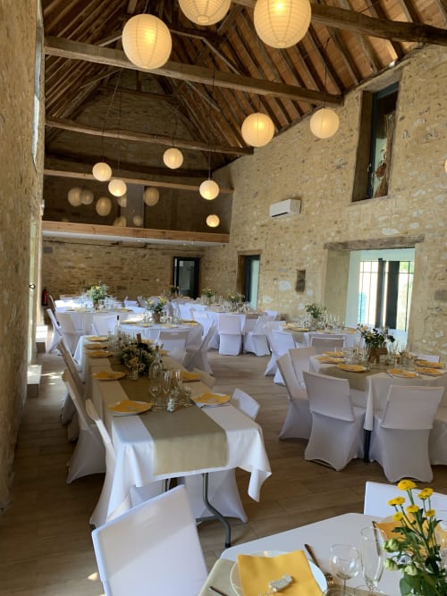 Élégante salle de banquet au Château de Baylac avec tables longues, chaises blanches, serviettes jaunes, lanternes suspendues et poutres apparentes
