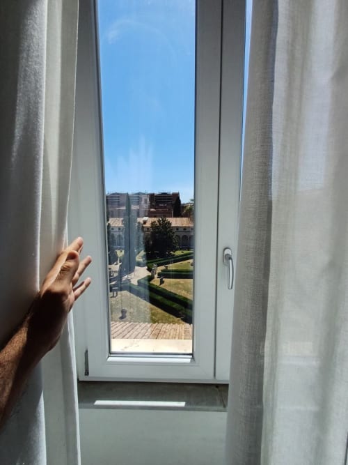 Mano che apre tende bianche per rivelare la vista del giardino della Michelangelo Suite Roma con edifici e cortile