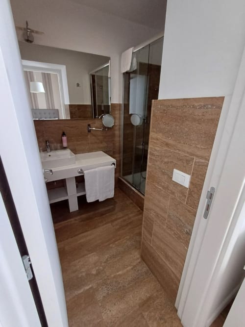Bagno moderno nella suite dell'hotel Roma con piastrelle marroni, doccia aperta, lavabo con piedistallo e asciugamani bianchi
