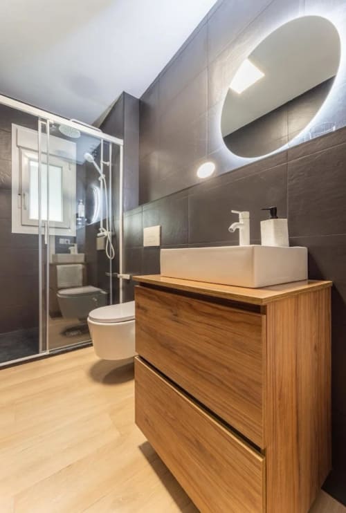Baño moderno en apartamento Gestyp-AT Mérida con mueble de madera, lavabo tipo vasija, espejo ovalado, paredes con azulejos oscuros y ducha de cristal