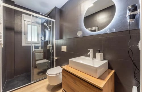 Baño moderno en apartamento Gestyp-AT, Mérida con azulejos oscuros, lavabo tipo recipiente, espejo circular y ducha con cerramiento de vidrio