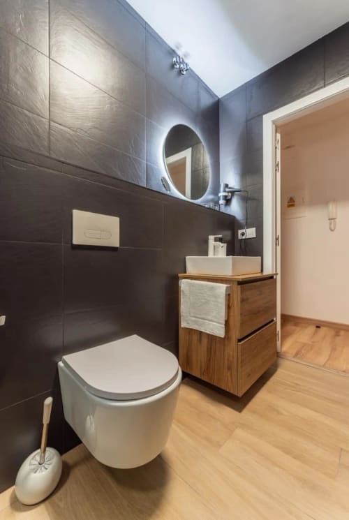 Baño moderno del apartamento Gestyp-AT en Mérida con azulejos negros, espejo ovalado iluminado, vanidad de madera e inodoro blanco