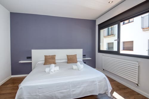 Modern bedroom in Calella apartment with purple accent wall, white bed, brown pillows, wooden floor, and large window