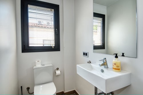 Modern bathroom in Calella apartment with white sink, toilet, mirror, window with blinds, and amenities dispenser