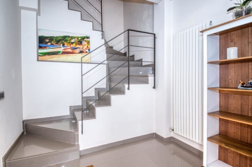 Modern staircase in Calella duplex apartment with glass railing, white walls, wooden shelving, and beach artwork at Finques