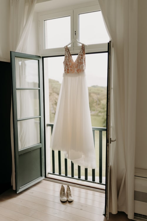 Élégante robe de mariage aux perles corail suspendue dans une pièce ensoleillée du Château de Baylac, avec chaussures en dessous