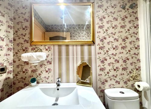 Baño moderno en apartamento de Baiona con lavamanos blanco, inodoro, espejo iluminado y papel tapiz floral morado