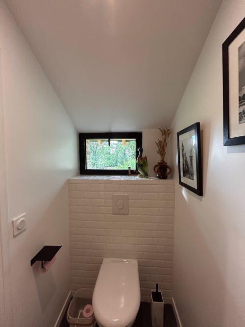 Salle de bain moderne du Domaine de Blanvatel avec toilettes blanches, carrelage, décoration encadrée et fenêtre sur la verdure