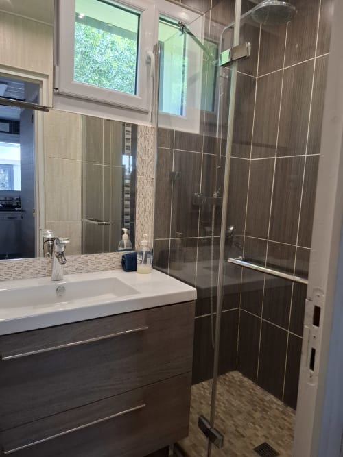 Salle de bain moderne du gîte Accolade à Châteaudun avec meuble vasque en bois foncé, douche vitrée et vue sur la rivière