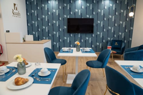 Sala colazione Ruah Pompei con carta da parati a motivi blu, tavoli in legno, sedie blu, coperti e pasticcini