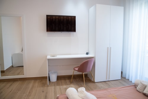 Modern hotel room at Ruah Pompei in Pompei with desk, pink chair, wall-mounted TV, wardrobe, and wooden flooring