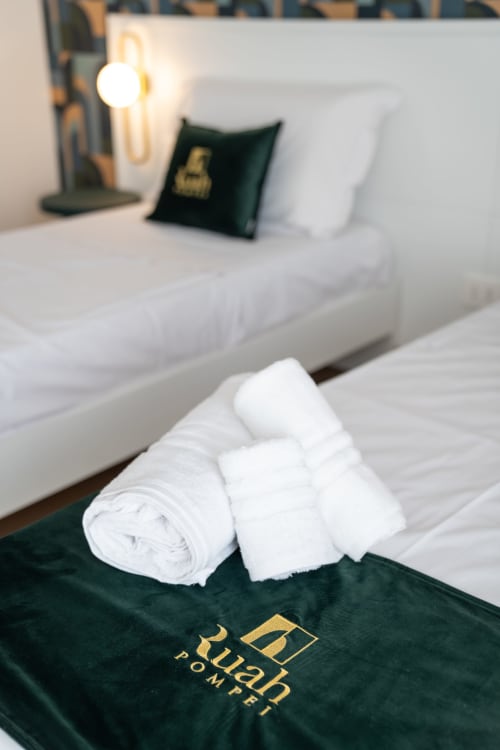 White rolled towels on green Ruah Pompei branded cloth in double bedroom with white bedding and desk lamp