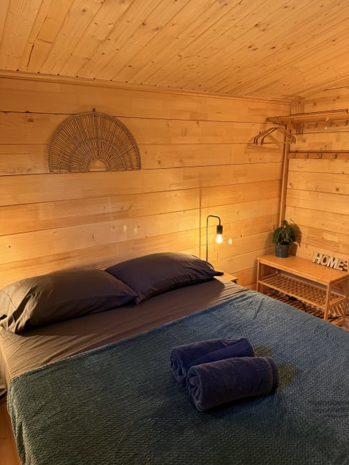Accogliente camera da letto in una baita di legno con letto, cuscini scuri, asciugamani arrotolati, lampada a sospensione, arte murale tessuta e pianta in vaso