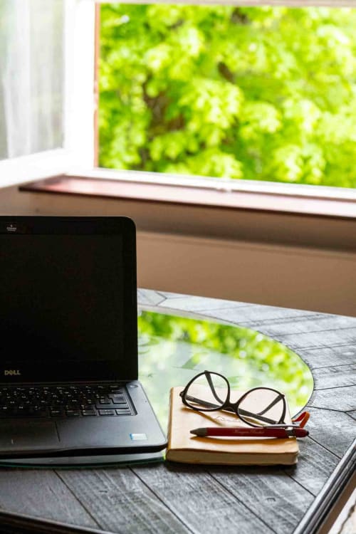 Bureau lumineux avec ordinateur, carnet et lunettes près fenêtre verdoyante, maison d’hôtes à Evry, Essonne, France, calme, inspiration