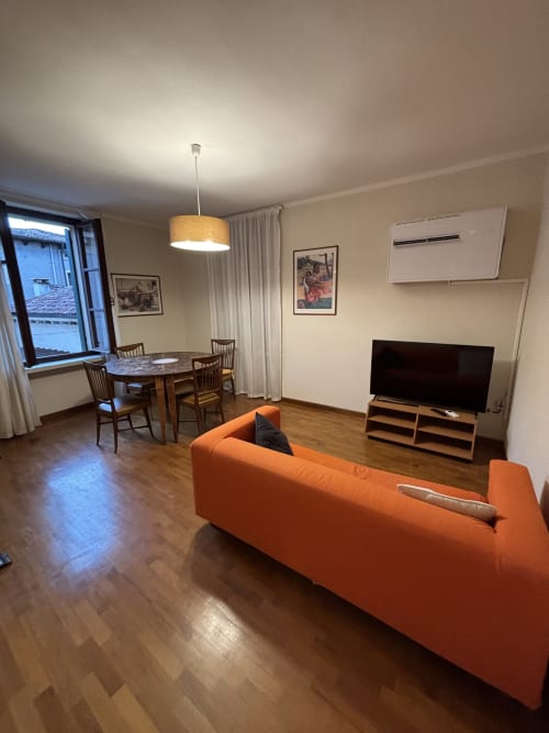 Soggiorno moderno in appartamento a Dimore Verona con divano arancione, TV, zona pranzo e vista sulla città dalle finestre