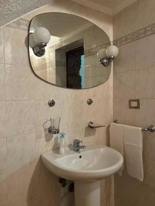 Bagno moderno della Residenza San Jacopo a Spello con lavabo a colonna, specchio rotondo con applique e piastrelle beige