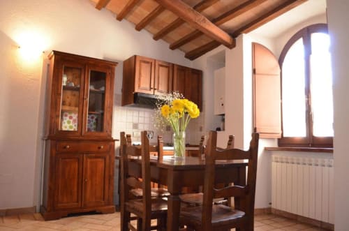 Rustica zona pranzo nell'appartamento di Spello con tavolo in legno, sedie, fiori gialli, travi a vista e finestra ad arco