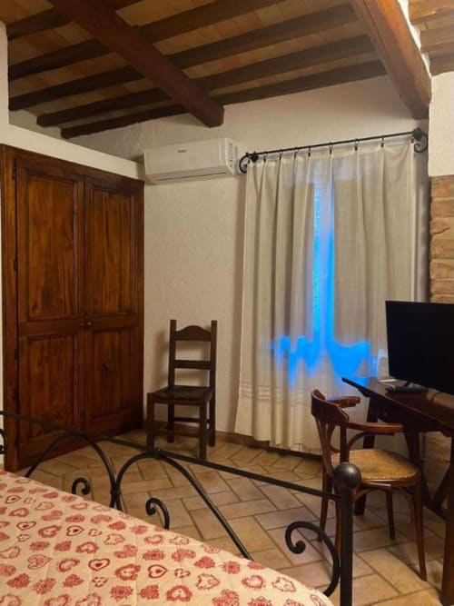 Camera B&B a Spello con travi a vista, armadio in legno, sedia, TV, tende color crema e pavimento in ceramica
