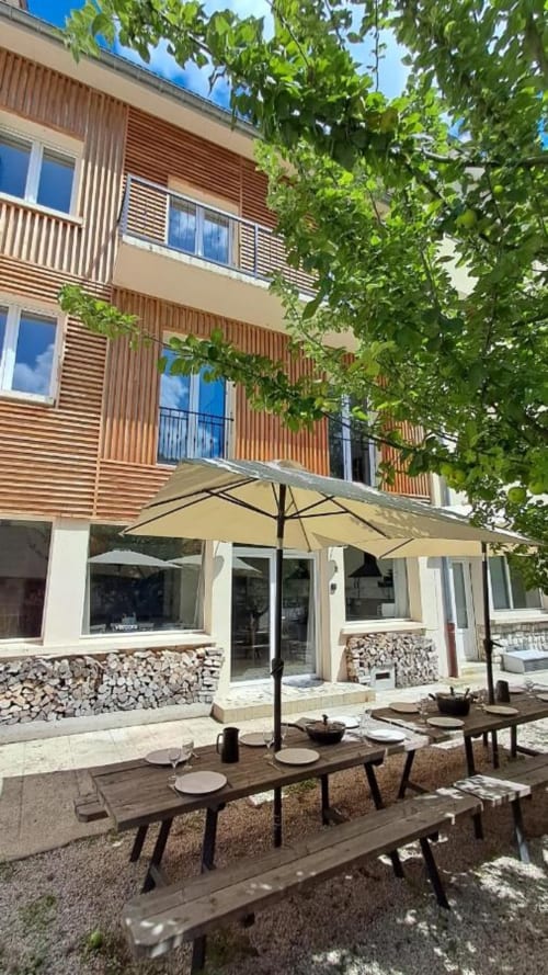 Cour de la Maison de Sophie avec tables en bois, parasols crème et façade aux lames de bois à Rencurel