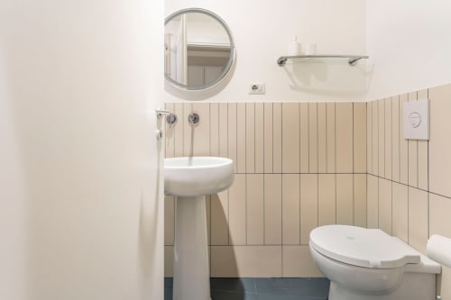 Bagno moderno dell'appartamento Art House Rome con specchio rotondo, lavabo a parete, wc e piastrelle beige.