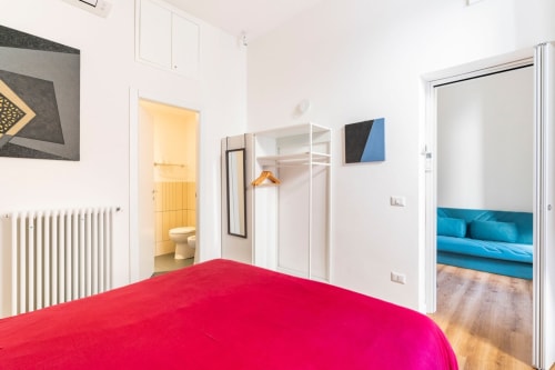 Camera luminosa con letto rosso, guardaroba bianco, bagno en-suite e divano blu nella stanza adiacente dell'appartamento Art House Roma