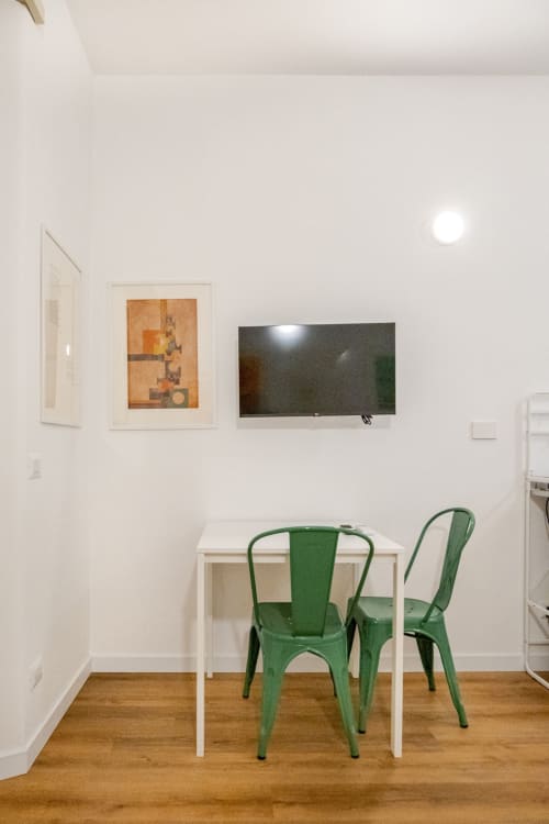 Moderno spazio di lavoro in camera d'hotel a Roma con tavolo bianco, sedie in metallo verde, TV a parete e quadri decorativi