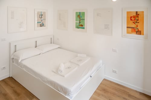 Camera da letto minimalista moderna con letto bianco, pavimentazione in legno e arte astratta incorniciata nell'appartamento Art House Roma.