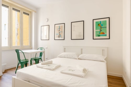Camera luminosa dell'Art House Roma con letto bianco, sedie verdi, scrivania, finestre e opere d'arte incorniciate