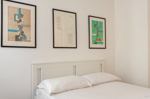 Camera moderna presso Art House Roma con letto bianco, due cuscini e tre opere d'arte astratta incorniciate su parete beige
