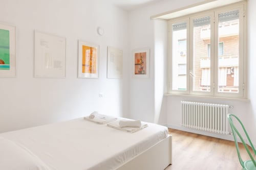 Camera luminosa in Art House Rome con letto bianco, opere d'arte incorniciate, radiatore e ampie finestre
