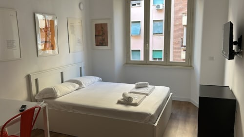Camera da letto luminosa e moderna all'Art House Roma con letto bianco, cuscini, quadri incorniciati, ampia finestra con vista su edificio in mattoni, rosso