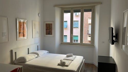 Camera luminosa all'Art House Roma con letto matrimoniale, biancheria bianca, quadri incorniciati e ampie finestre affacciate su edifici in mattoni