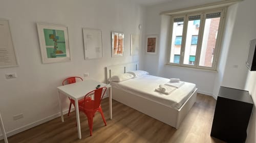Camera da letto moderna ad Art House Roma con letto bianco, scrivania e sedia rossa, opere d'arte incorniciate e ampie finestre con vista panoramica