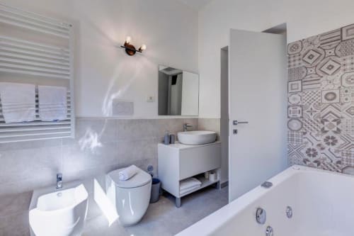 Modern bathroom suite at Palazzo Belvedere with white vanity, toilet, bathtub, patterned tile accent wall, and natural light