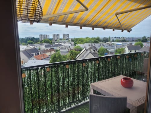 Balcon avec store jaune, garde-corps couvert de lierre vert, bol rouge, skyline de la ville à Val'Déco, Les Ponts de Cé
