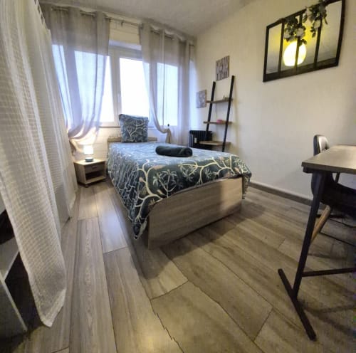 Chambre moderne à Val'Déco Conciergerie avec lit simple, bureau, parquet, grandes fenêtres et vue sur la ville aux Ponts de